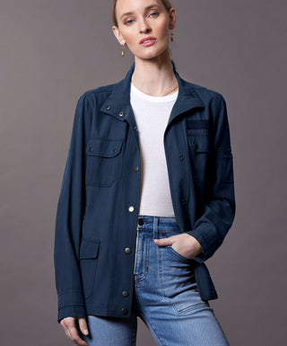 A woman modeling a dark navy blue utility jacket over a white ribbed top and blue jeans, photographed against a solid dark gray studio background.