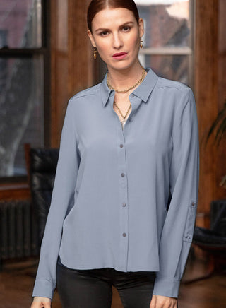 A woman wearing a light blue long-sleeve button-up blouse with a collar and dark buttons, paired with black pants, standing in a dimly lit room with wood paneling.